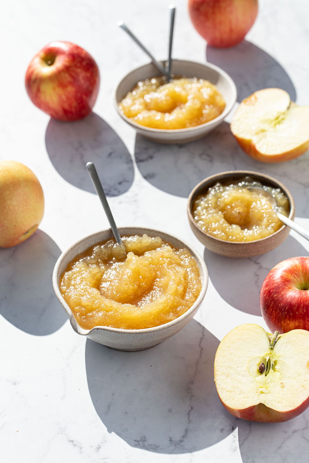 Multiple different sized bowls full of homemade applesauce on a marble background, with a few whole and halved apples scattered around.