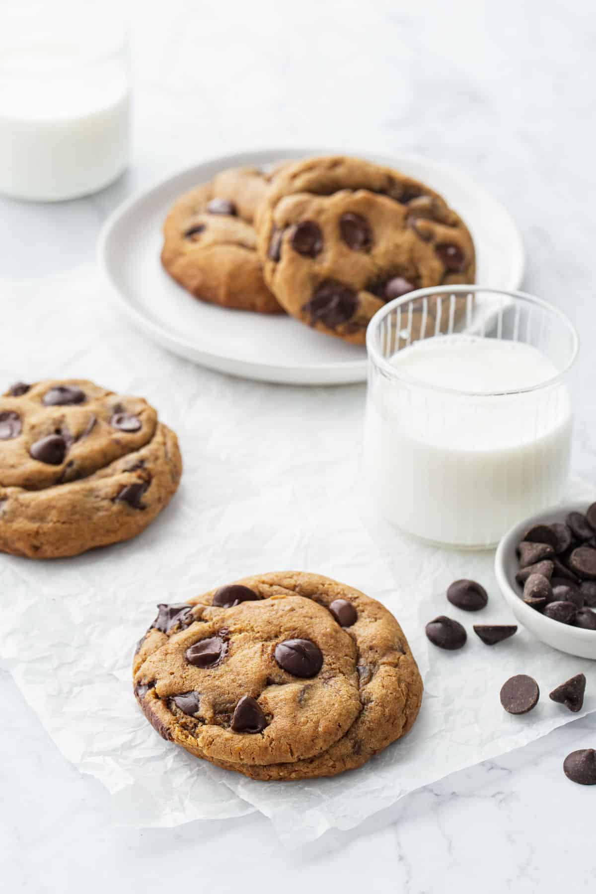 Three extra large Brown Butter Chocolate Chip Cookies on a white background, with two small glasses of milk and a bowl of chocolate chips on the side.